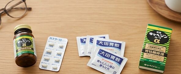 木製のテーブルの上に並べられた各種市販胃腸薬（錠剤シート、粉末包、ボトル）と、「胃酸」「胃もたれ」「胃粘膜修復」と書かれた症状別選び方の解説カードの並び
